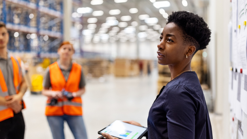 Supervisor standing by a whiteboard with a digital tablet discussing dispatch plan with workers.  Team of workers having meeting in a distribution warehouse.